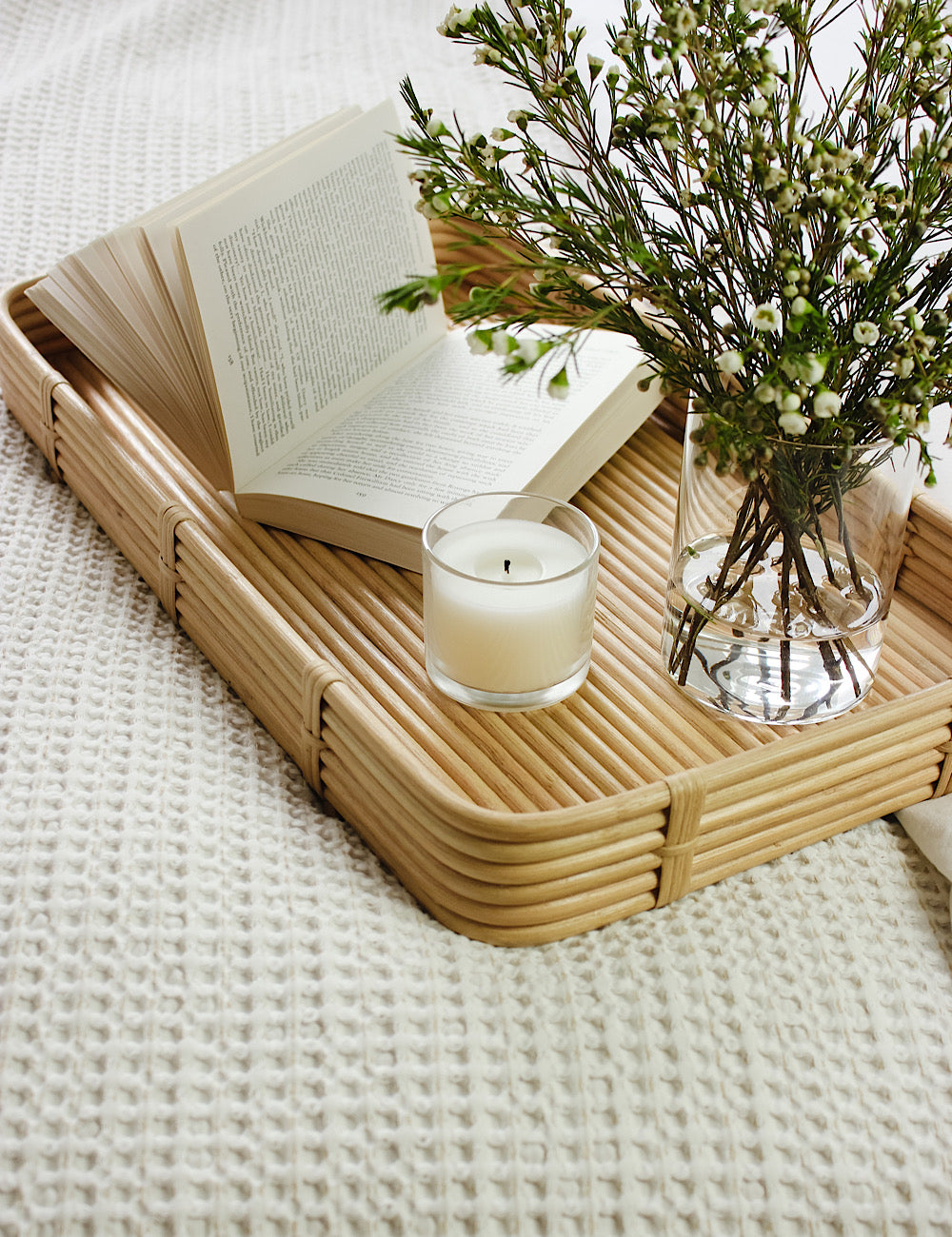 Styled shot of a rectangular bamboo tray on a bed, accessorised with flowers, a candle, and an open book.
