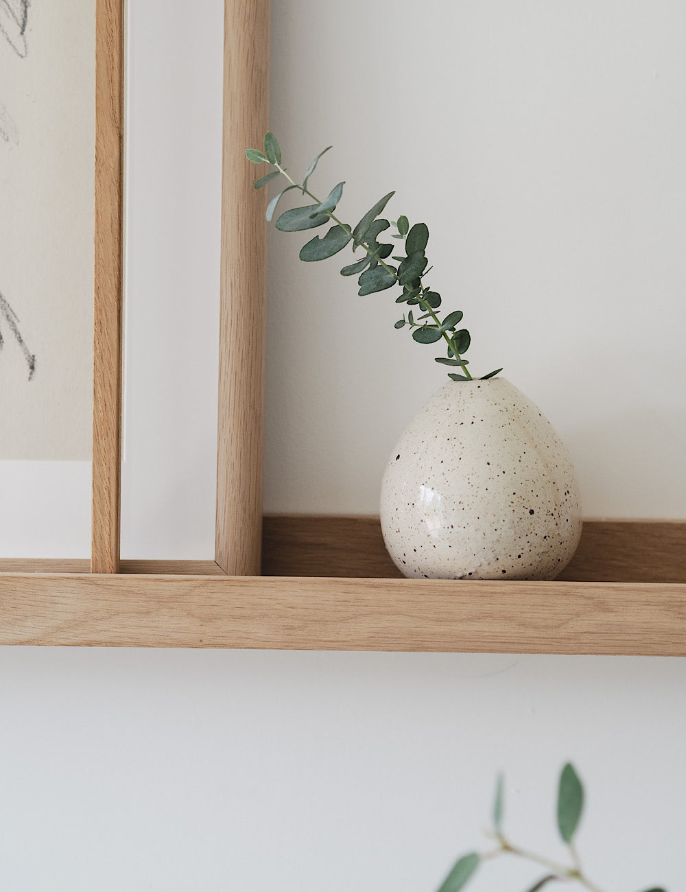 Styled image shows the Juno, a small ceramic vase with a rugged texture and tear drop shape on a shelf with a green eucalyptus stem inside it. 