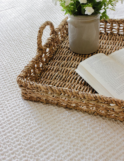 Side shot of a rattan tray styled on a bed with flowers and a book.