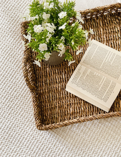 Top shot of a square rattan tray styled on a bed with flowers and a book.